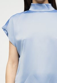 Γυναίκα που φοράει γαλάζιο σατέν μπλουζάκι με κοντά, φαρδιά μανίκια και ψηλό, διπλωμένο λαιμόκοψη, πάνω σε λευκό φόντο.