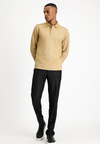 Beige Langarm-Polohemd mit Kragen und einer Knopfleiste mit drei Knöpfen, kombiniert mit schwarzen Hosen und schwarzen Sneakers. Sichtbare Details aus strukturiertem Stoff.