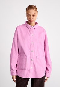 Mladá žena s vrkôčkami, oblečená v ružovej kordovej bundě na gombíky a tmavých nohaviciach, stojí pred jednoduchým svetlým pozadím.