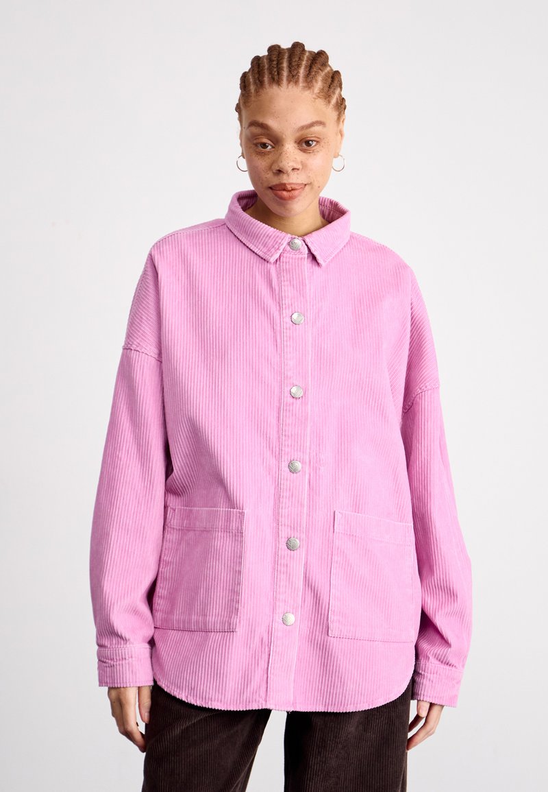 Mladá žena s vrkôčkami, oblečená v ružovej kordovej bundě na gombíky a tmavých nohaviciach, stojí pred jednoduchým svetlým pozadím.