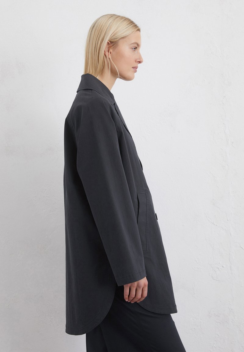 Czarny długi płaszcz w odcieniu ciemnoszarym, z kołnierzykiem, bocznymi kieszeniami i zaokrąglonym dołem. Wykonany z gładkiego materiału, charakteryzujący się luźnym krojem i minimalistycznym designem.