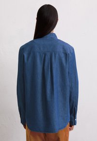 Blauwe denim shirt met kraag, lange mouwen en een detail met een rugplooi. Soepele textuur, gebogen zoom en met knopen sluitende manchetten.