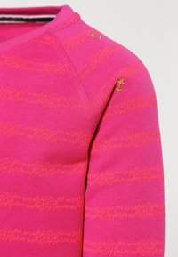 Fuksjowa bluzka z długim rękawem z nadrukiem tekstowym w kolorze pomarańczowym. Charakteryzuje się teksturowanym wykończeniem i złotym akcentem w kształcie kotwicy na ramieniu.