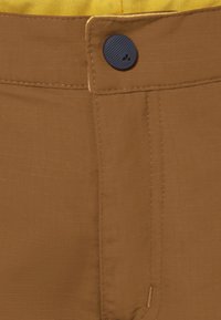 Pantaloni di stoffa marrone con una superficie testurizzata, dotati di un bottone blu e di un tessuto giallo visibile sopra. I punti di cucitura sono ordinati e strutturati.