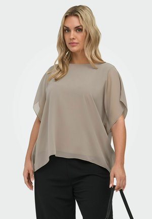 Lyshåret kvinde iført løs taupebluse og sorte bukser, der holder en sort rem mod en ensartet lys baggrund.