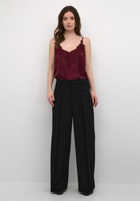 Camisola di raso bordeaux con rifiniture in pizzo; abbinata a pantaloni neri plissettati a vita alta. La texture morbida e la vestibilità ampia mettono in risalto la silhouette.