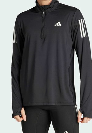 Schwarzes langärmeliges Sportoberteil mit Viertel-Reißverschluss, Adidas-Logo auf der Brust, drei weißen Streifen an den Ärmeln, getragen mit schwarzen Leggings.