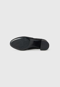 Scarpa nera con tacco basso mostrata dal fondo, con un motivo di presa testurizzato sulla parte anteriore e un'area del tacco liscia su uno sfondo neutro.