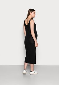 Vestido preto, sem mangas, com decote em V nas costas, silhueta ajustada e comprimento até o tornozelo, combinado com sapatos brancos com detalhes pretos.