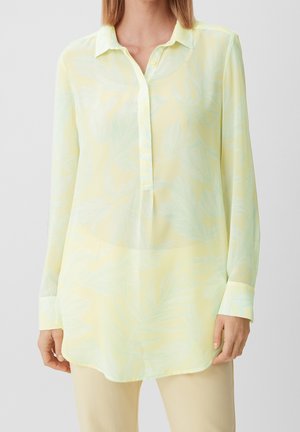 Kobieta ubrana w długą, jasnżółtą, półprzezroczystą bluzkę z delikatnym kwiatowym wzorem i kołnierzykiem, w połączeniu z beżowymi spodniami.