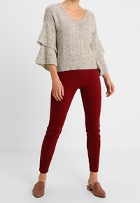 Pull gris clair en tricot avec col en V et manches évasées, accompagné d'un pantalon en velours côtelé bordeaux ajusté et de chaussures marron sans lacets.