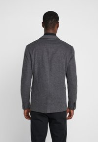 Blazer de lana gris con un diseño estructurado, abertura simple y solapas con muescas. Presenta dos botones y un tejido texturizado. Corte casual, con una camisa negra debajo.