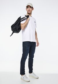 T-shirt bianco, jeans blu scuro, sneakers bianche. Zaino nero con spallacci regolabili. Cappellino beige chiaro. Modello che posa su uno sfondo bianco.