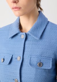 Modrá texturovaná bunda s klasickým límcem, dvěma předními kapsami se stříbrnými knoflíky a strukturovaným designem.