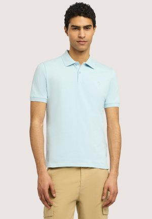 Uomo con capelli ricci che indossa una polo azzurra a maniche corte e pantaloni cargo beige, in piedi davanti a uno sfondo semplice.