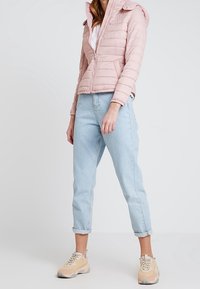 Lys rosa dunjakke med hætte, lynlås-lukning og ribbede detaljer, parret med lyseblå opsmøjede jeans og beige sneakers.