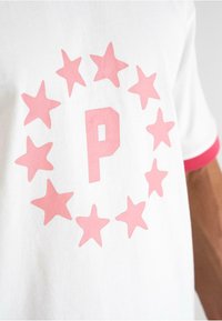 Baltā kokvilnas T-krekls ar apļveida rozā zvaigžņu dizainu centrā ar lielu rozā burtu "P". Piedurknes ir ar rozā apdari.