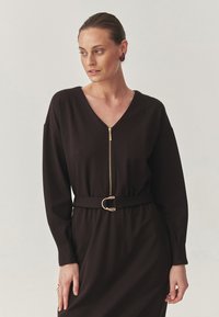 Zwarte langemouwen jurk met V-hals en gouden rits, nauwsluitend om de taille met een bijpassende riem met een ronde gesp.