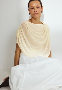 Beżowa bluzka w lekkim materiale o miękkiej fakturze, zestawiona z zwiewną białą spódnicą. Zauważalne kolczyki dodają kontrastu.