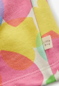 Kleurige stof met abstracte vormen in roze, geel, blauw en groen. Een wit label aan de rand met de tekst "Little Bird." Getextureerd katoen.