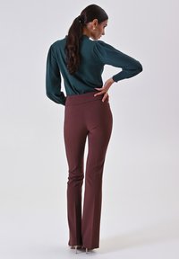 Pantaloni a palazzo marroni con vita alta, realizzati in un tessuto liscio. Abbinati a una blouse manica lunga verde scuro. Lo sfondo è semplice.