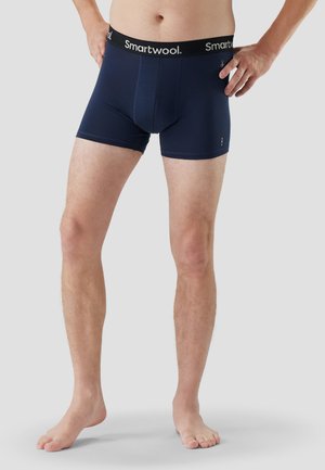 Man draagt marineblauwe Smartwool boxer shorts, staat op blote voeten op een lichtgrijze achtergrond, met handen op de heupen.