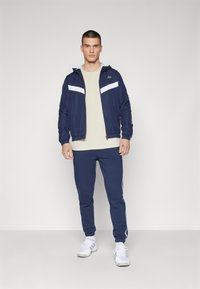 Giacca a track navy con strisce bianche e cappuccio, abbinata a pantaloni tuta navy e scarpe da ginnastica bianche. Abbigliamento sportivo casual.