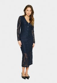 Navy lace dress with long sleeves, V-neck, and button closure. Textured floral pattern and semi-transparent lower section. High slit design.