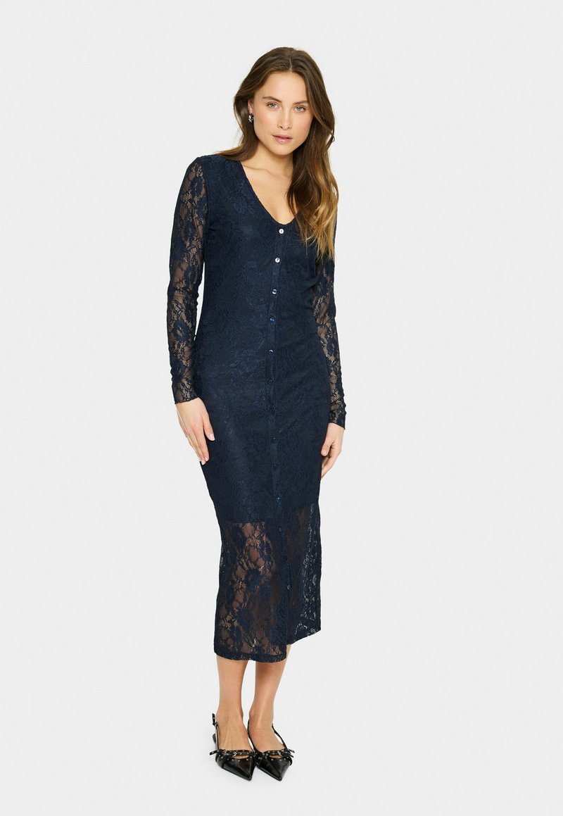 Navy lace dress with long sleeves, V-neck, and button closure. Textured floral pattern and semi-transparent lower section. High slit design.