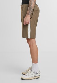 Shorts kaki avec une bande latérale blanche, ornés d'un détail chaîne à la taille, confectionnés dans un tissu l éger. Portés avec des chaussures de sport blanches.