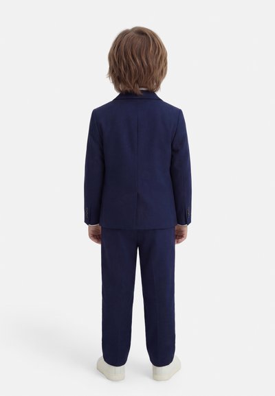Παιδί με μεσαίου μήκους καστανά μαλλιά, που στέκεται με την πλάτη προς το φακό, φοράει μπλε σκούρο κοστούμι και λευκά αθλητικά παπούτσια, με φόντο ένα άσπρο τοίχο.