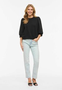 Femme debout souriante, portant un haut noir à manches bouffantes, un jean bleu clair, et des sandales à talons ouvertes noires, main dans la poche.