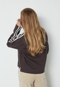 Mujer con cabello largo y rubio, vistiendo un sudadera marrón oscuro con rayas blancas en las mangas y pantalones beige, de frente a una pared blanca.