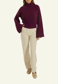 Frau trägt einen gerippten, lila Rollkragenpullover mit weiten Ärmeln, cremefarbene, gerade Hosen und beigefarbene Slipper und steht mit einer Hand auf der Hüfte.