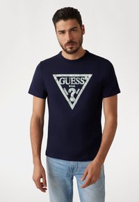 Navy cotton t-shirt with a triangular "GUESS" logo in white and silver, featuring intricate patterns and a crew neckline.