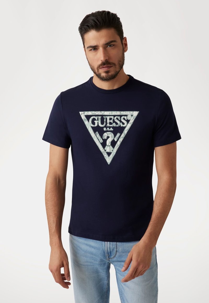 Camiseta de algodón azul marino con un logo triangular de "GUESS" en blanco y plata, que presenta patrones intrincados y un escote de cuello redondo.
