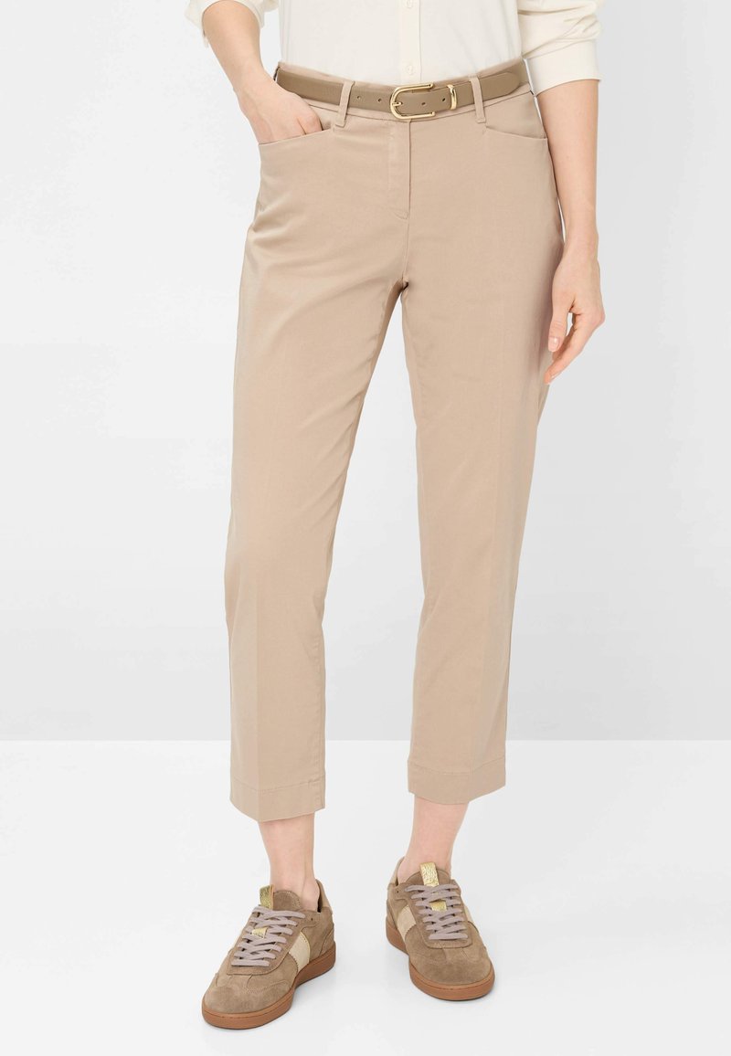 Persoon draagt beige cropped broek met riem, gecombineerd met bruine suède sneakers met lichtbeige accenten.