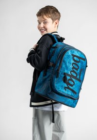 Modrý batoh s vzorem a černými akcenty, výrazným logem, obdélníkovým tvarem, polstrovanými popruhy a několika zipovými kapsami.