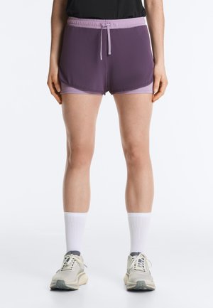 Lábak, amelyek réteges lila sportnadrágot, fehér középmagas zoknit és világos színű futócipőt viselnek, egyszerű fehér háttér előtt.