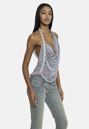 Woman wearing a sleeveless gray draped halter top with buttons and faded blue jeans, standing against a plain background.