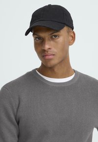 Fekete baseballsapka ívelt karimával, pamutból készült, egy szürke texturált pulóver felett viselve, amelynek kerek nyaka és fehér alsó pólója van.