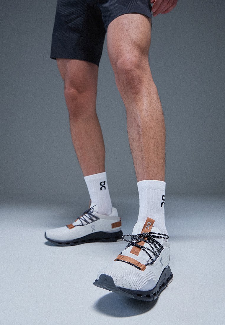 Weiße Sportschuhe mit schwarzen Schnürsenkeln, strukturierten grauen Sohlen und orangefarbenen Akzenten am Obermaterial. Kombiniert mit knielangen weißen Socken mit Logo.
