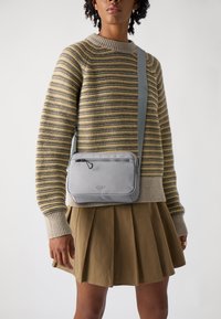 Borsa a tracolla grigia rettangolare con chiusura a zip, abbinata a un maglione a righe e una gonna beige plissettata, posizionata su uno sfondo semplice.