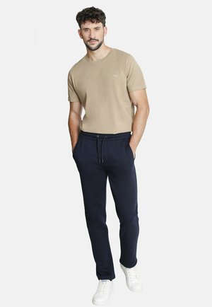 Camiseta beige de manga corta, combinada con joggers azul marino que tienen una cintura elástica y cordón. El modelo lleva zapatillas blancas.