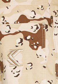 Beige och brun ökenkamouflage tyg med en sydd ficka och en metallisk broderad logotyp på nedre vänstra sidan.