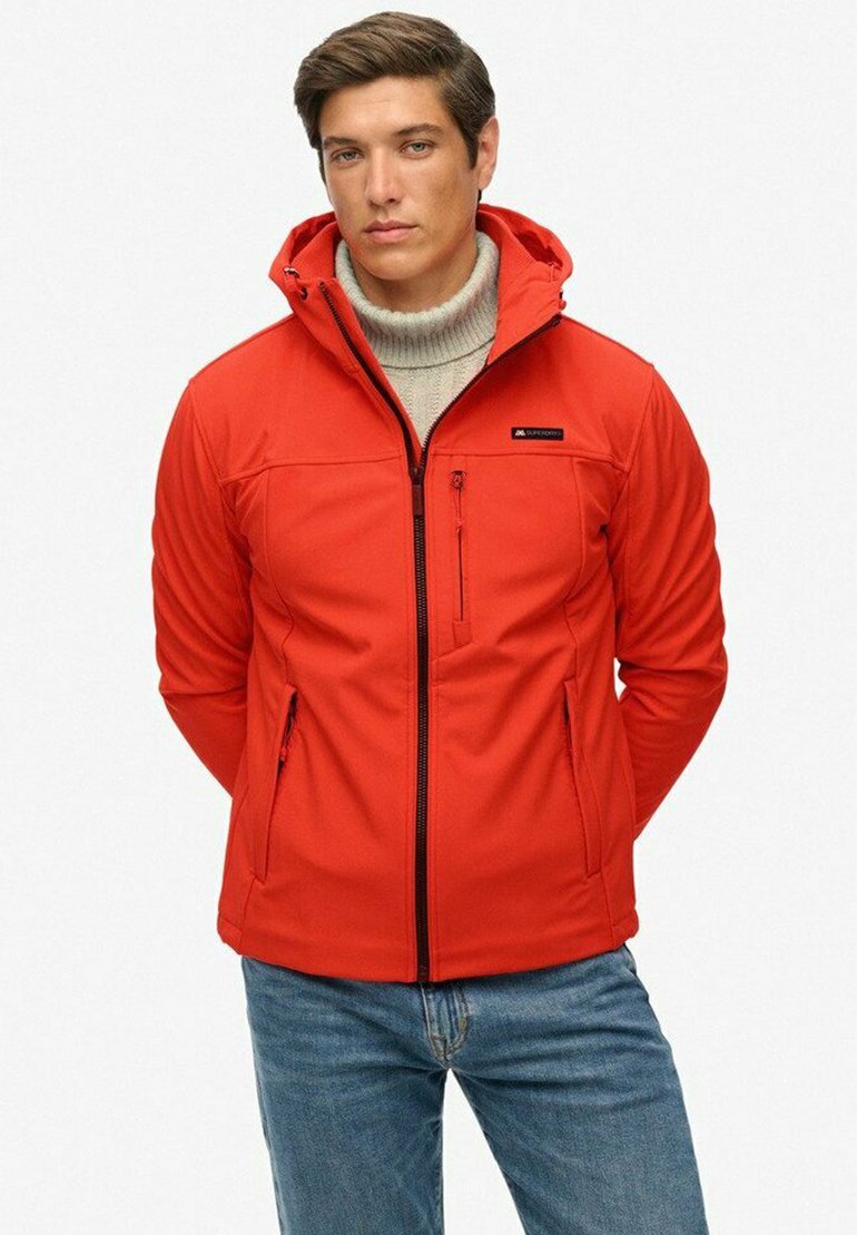 Veste softshell rouge avec fermeture éclair, capuche ajustable et poche poitrine zippée. Portée par-dessus un pull col roulé gris clair.