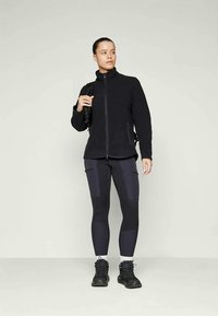 Svart zip-up jacka med sidofickor, parat med mörka leggings med lappar. Modellen bär svarta sneakers och håller en ryggsäck.