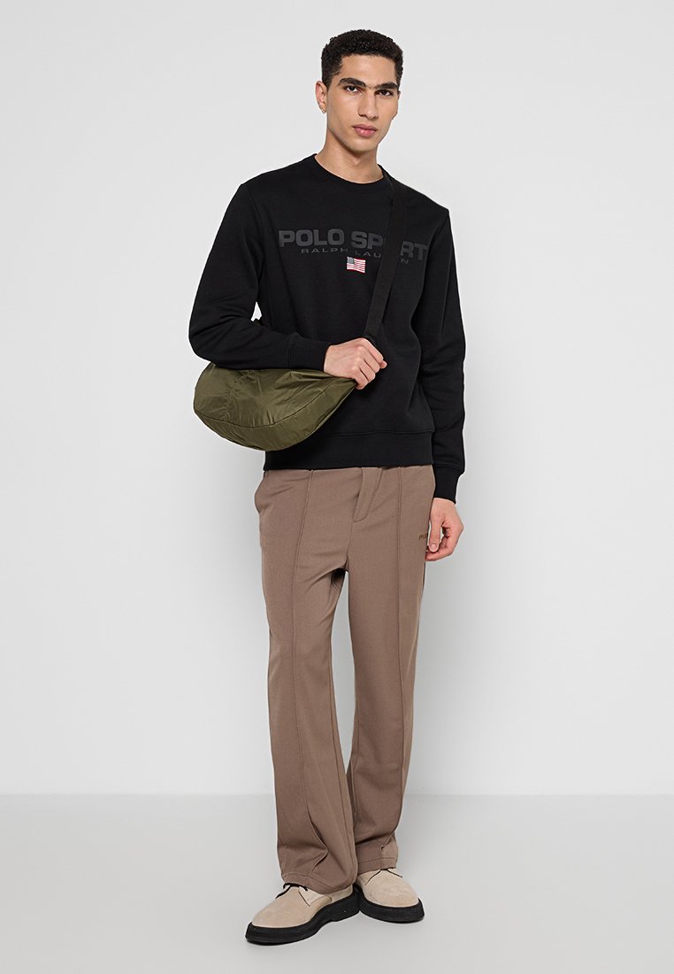 Felpa nera con logo "POLO SPORT", pantaloni larghi color sabbia e borsa a tracolla verde. Il modello è in piedi di fronte a uno sfondo neutro.