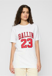 Baltos medvilnės marškinėliai su laisvu kirpimu, ant priekinės pusės ryškiai užrašyti raudoni blokiniai žodžiai „BALLIN“ ir skaičius „23“.}}