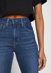 Hoch taillierte blaue Denim-Jeans mit einer glatten, dunklen Waschtextur, ausgestattet mit einem klassischen Knopf- und Reißverschluss sowie vorderen Taschen.